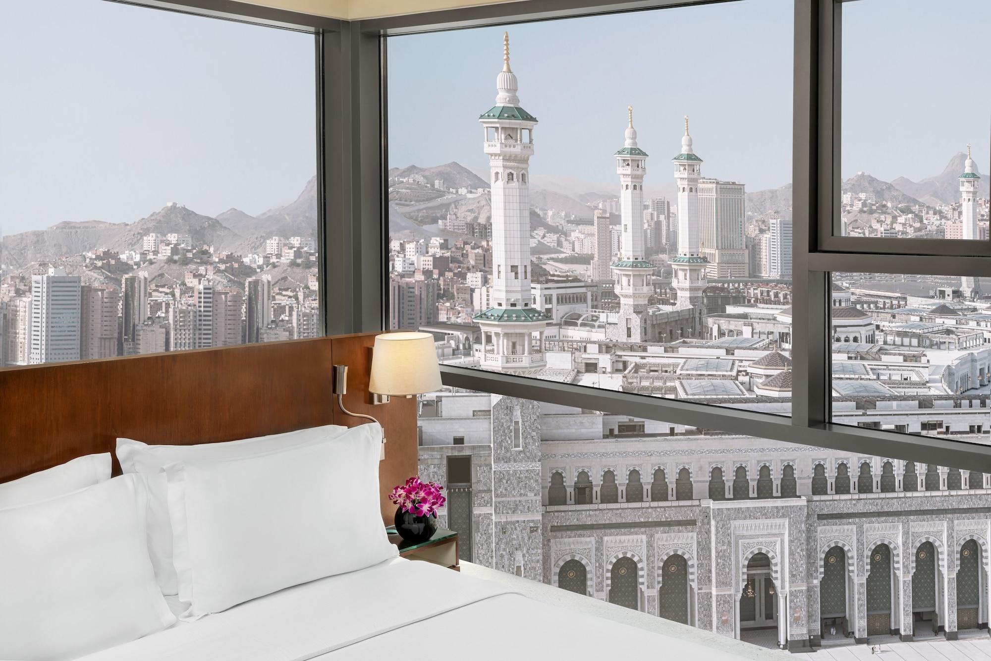 Hilton Suites Jabal Omar Makkah Meca Exterior foto The photo depicts a hotel room with a modern design. In the foreground, there is a neatly made bed with white linens and a bedside lamp. A small flower arrangement adds a touch of color to the room. Through large windows, a stunning view of a citysca