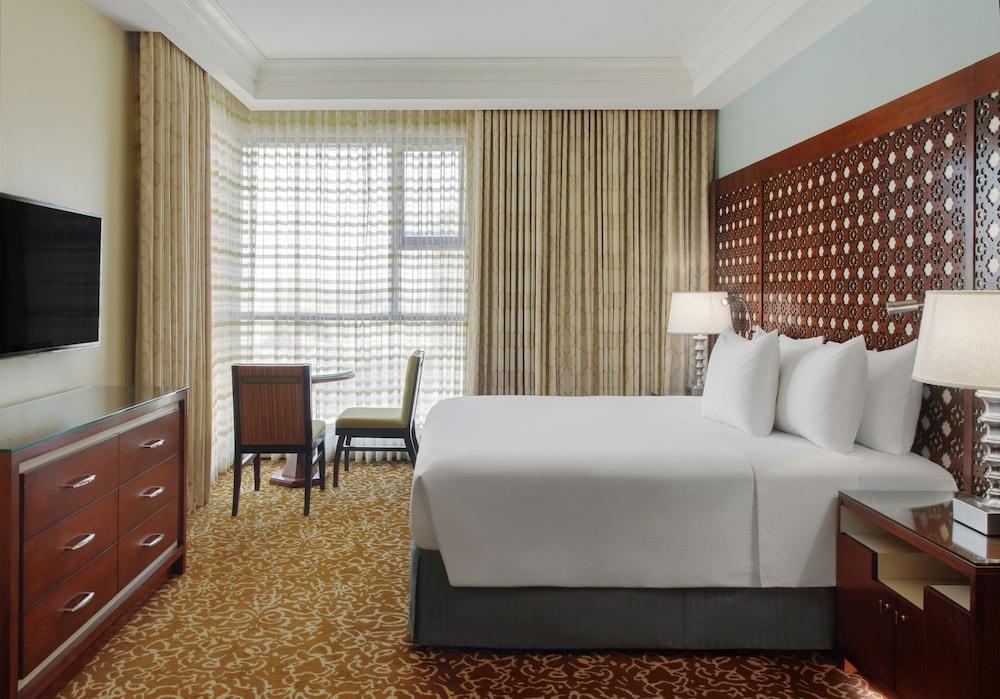 Hilton Suites Jabal Omar Makkah Meca Exterior foto The photo shows a hotel room featuring a large bed with white linens and several pillows. There is a wooden headboard with a decorative design. Alongside the bed, there are bedside lamps. A wooden dresser is visible, with a television mounted on the 