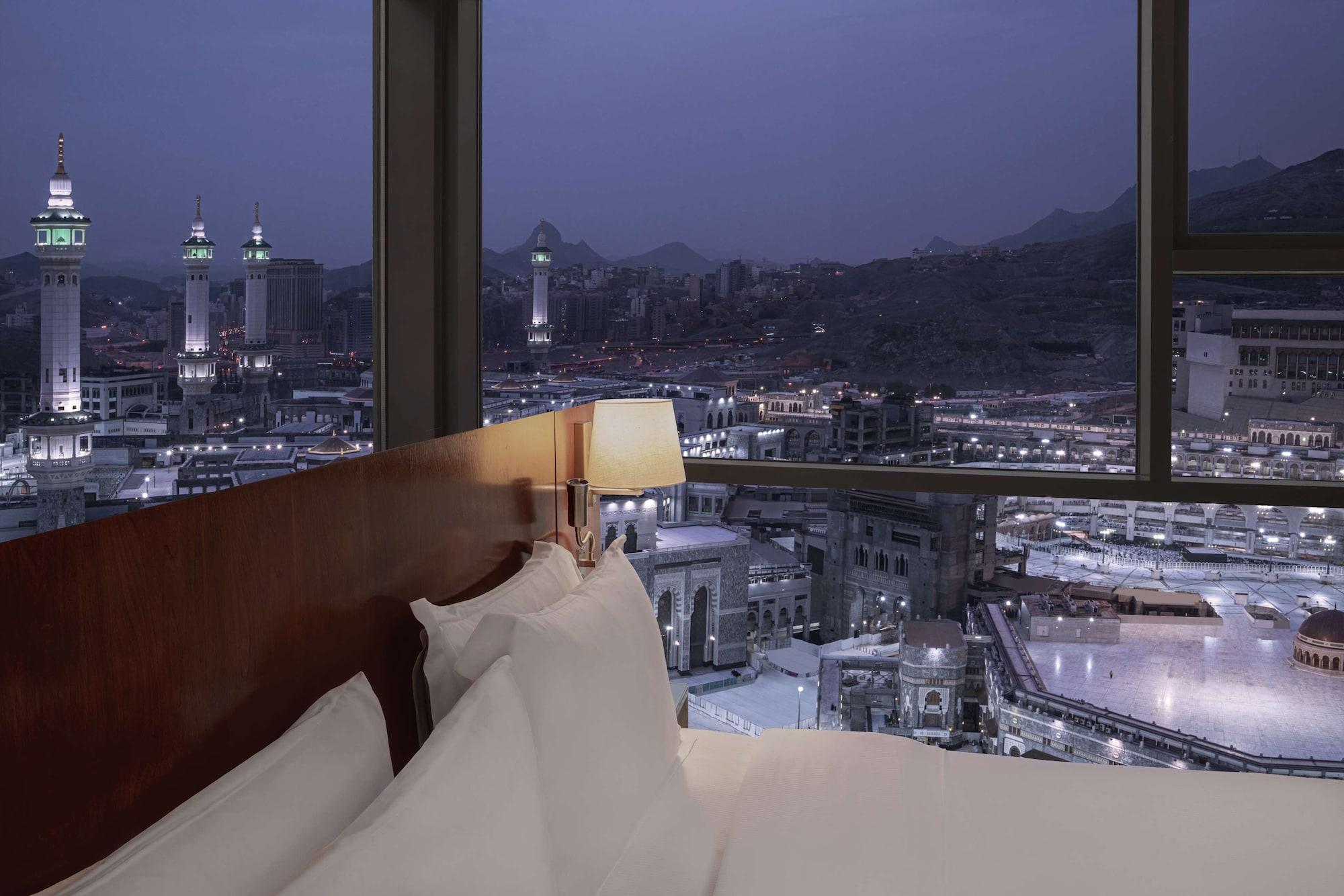 Hilton Suites Jabal Omar Makkah Meca Exterior foto The photo depicts a view from inside a hotel room at night. The foreground shows a bed with white pillows and linens. In the background, large windows offer a panoramic view of a cityscape, featuring notable minarets and buildings illuminated against