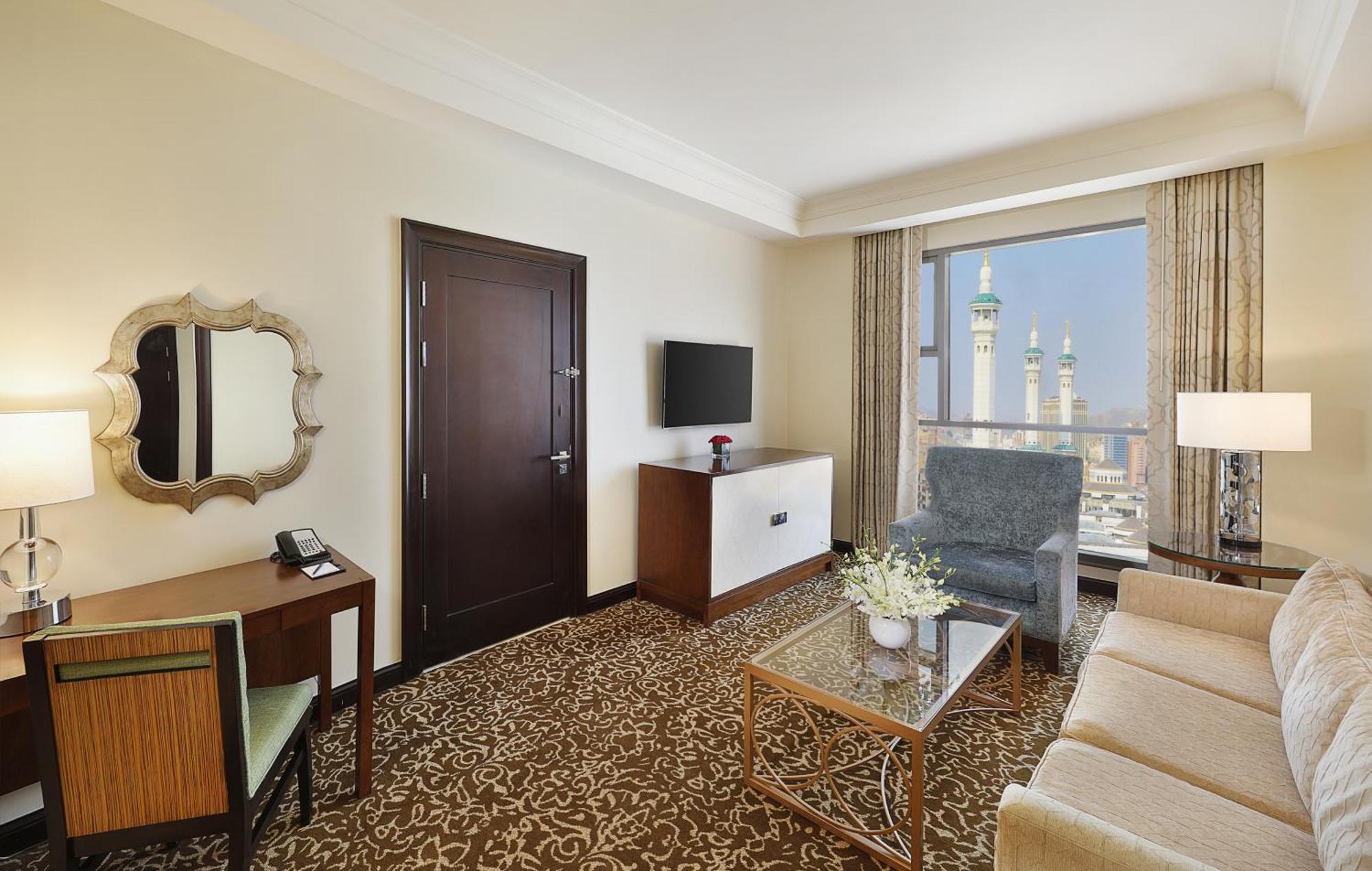 Hilton Suites Jabal Omar Makkah Meca Exterior foto The photo shows a hotel room with a modern and elegant design. It features a cozy seating area with a beige sofa and a blue armchair, complemented by a glass coffee table. There is a wooden desk with a mirror and a phone, and a television mounted on 
