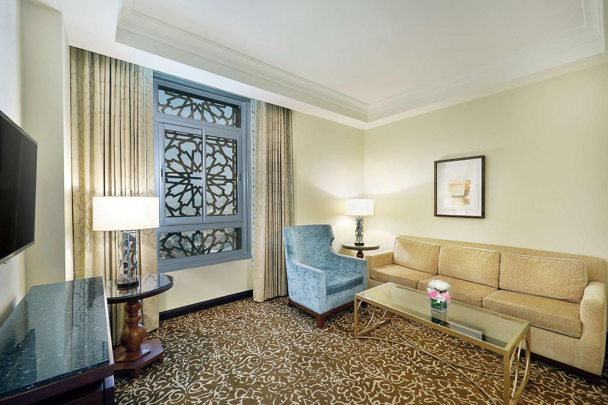 Hilton Suites Jabal Omar Makkah Meca Exterior foto The photo shows a well-decorated hotel room or suite. It features a light-colored wall with a decorative window that has an intricate design. There are two windows adorned with curtains. The furniture includes a beige couch, a blue upholstered armcha