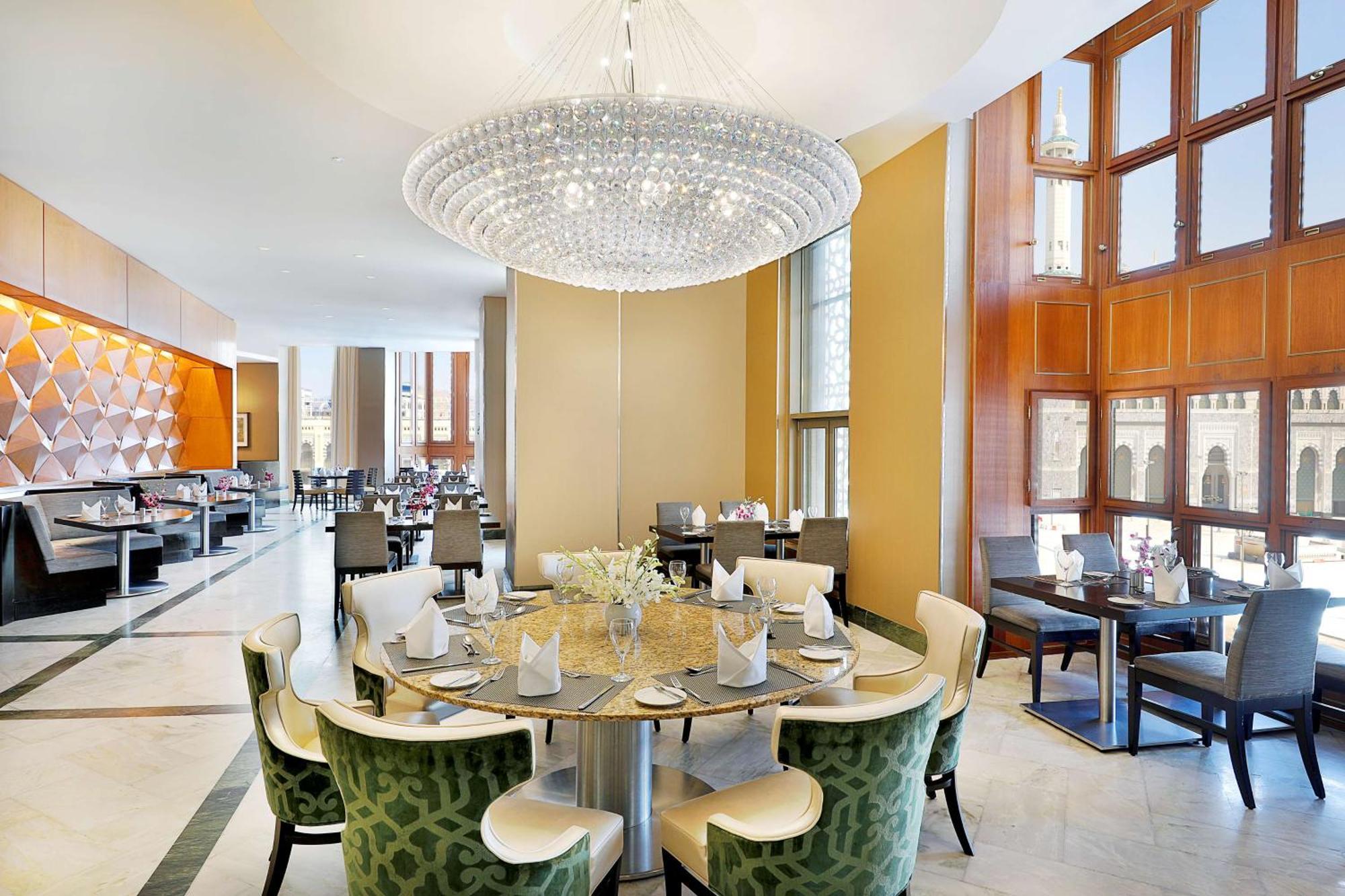 Hilton Suites Jabal Omar Makkah Meca Exterior foto The photo shows a modern restaurant interior. In the center, there's a round dining table with green upholstered chairs, elegantly arranged with plates and napkins. Above the table hangs a large crystal chandelier, providing bright, stylish lighting.