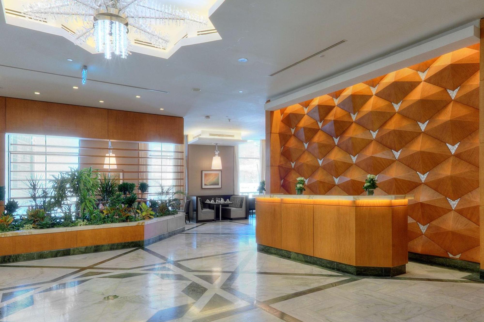 Hilton Suites Jabal Omar Makkah Meca Exterior foto The photo shows a hotel lobby area. On the right side, there is a reception desk made of wood, featuring a modern design. Behind the desk, there is a decorative wall with a unique geometric pattern in shades of orange and brown. To the left, there ar