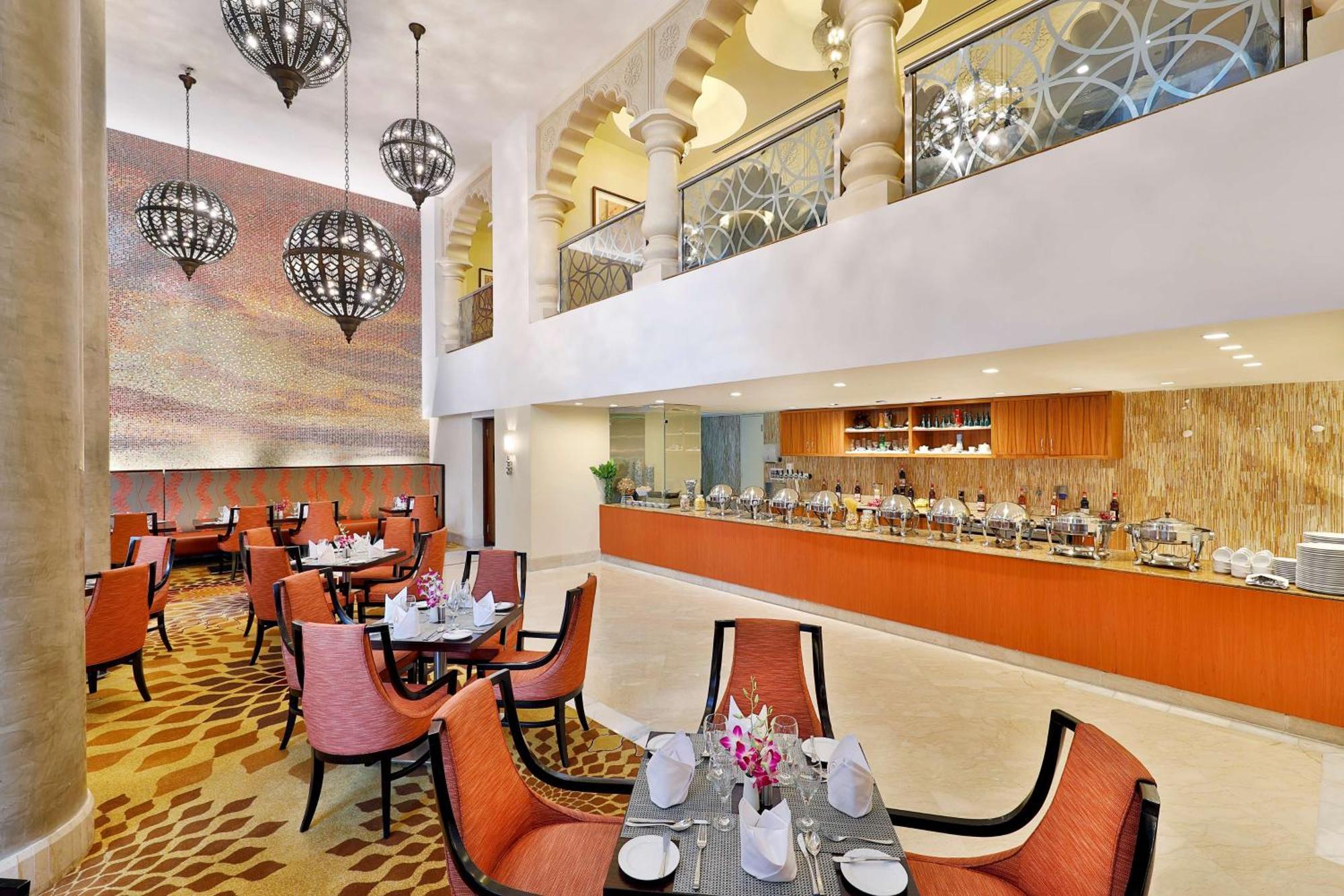 Hilton Suites Jabal Omar Makkah Meca Exterior foto The photo shows a modern restaurant interior with a spacious and elegant design. It features a dining area with several round tables surrounded by orange chairs. Each table is set with white tableware and adorned with small flower arrangements. 

In 