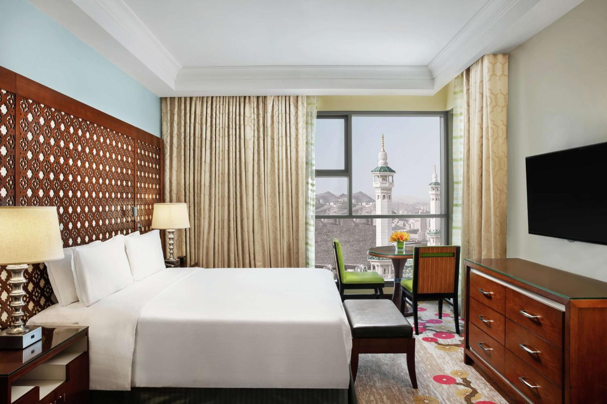 Hilton Suites Jabal Omar Makkah Meca Exterior foto The photo shows a well-furnished hotel room. It features a large bed with white bedding, a wooden headboard, and two bedside lamps. There’s a small seating area with a chair and a table near a window that provides a view of a mosque's minarets outsid