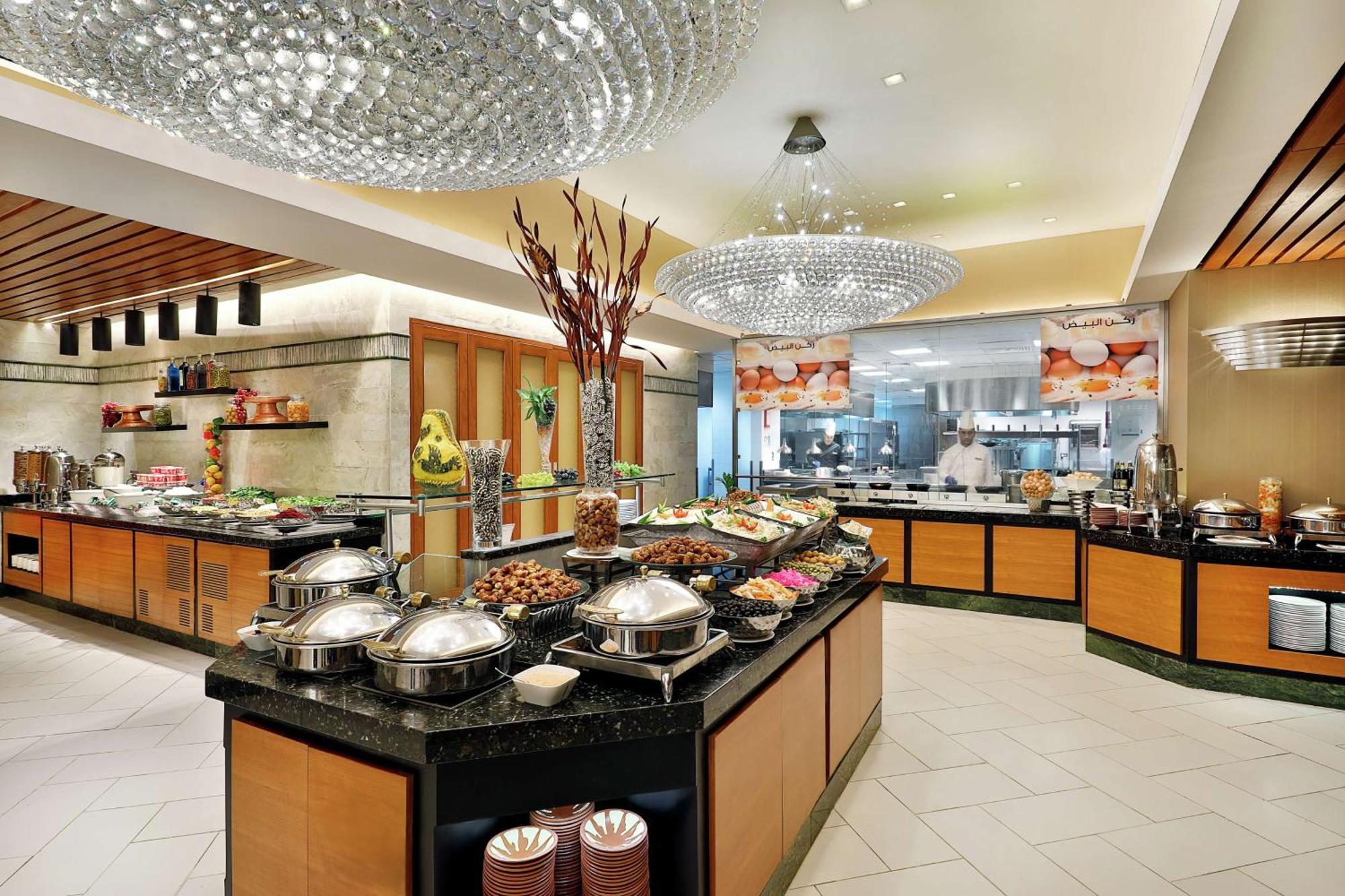 Hilton Suites Jabal Omar Makkah Meca Exterior foto The photo shows a spacious buffet area in a restaurant or hotel. The design features elegant decor, including bright chandeliers and polished surfaces. There are several buffet tables filled with various food items, displayed attractively. The tables