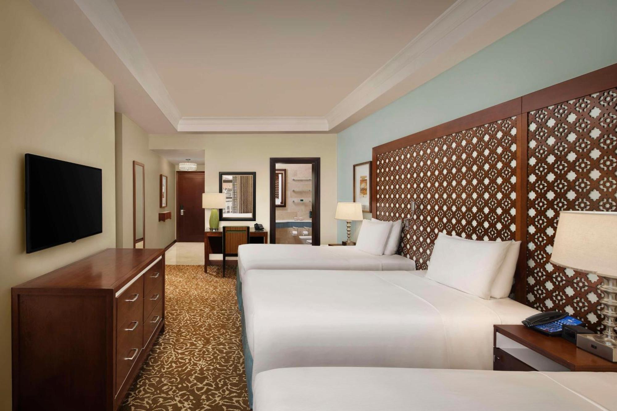 Hilton Suites Jabal Omar Makkah Meca Exterior foto The photo shows a hotel room featuring two beds with white linens. There is a decorative headboard behind each bed, made of wood with a patterned design. The room has a warm beige carpet and light blue walls, giving it a serene atmosphere. A wooden d