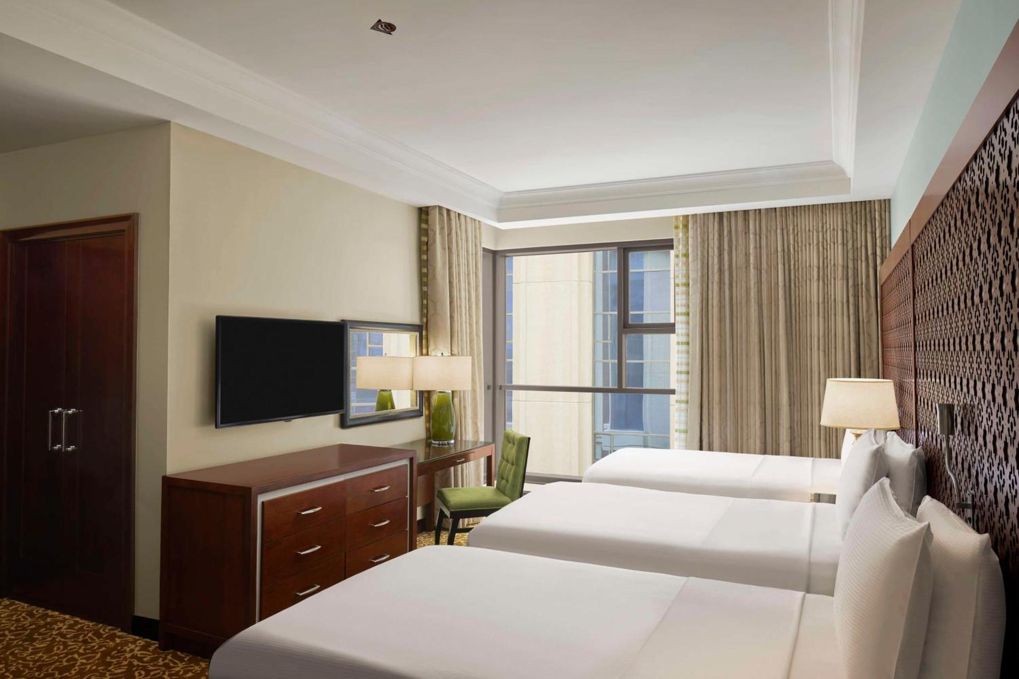Hilton Suites Jabal Omar Makkah Meca Exterior foto The photo shows a hotel room featuring two twin beds with white bedding. There's a wooden chest of drawers, a lamp, and a chair beside the window, which has light-colored curtains. A flat-screen TV is mounted on the wall across from the beds. The roo