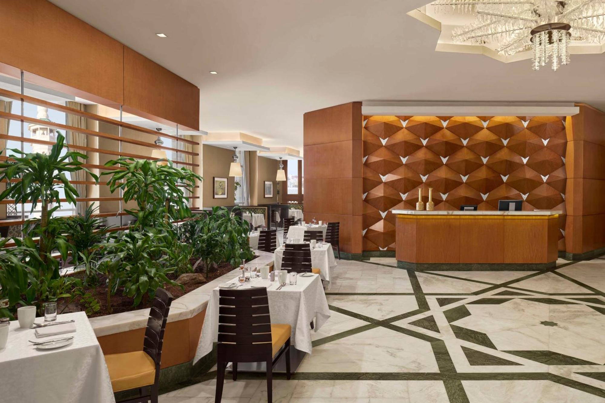 Hilton Suites Jabal Omar Makkah Meca Exterior foto The photo depicts an elegant restaurant interior. The space features a sleek design with wooden elements and a stylish reception area. To the left, there are neatly arranged dining tables with white tablecloths, each set with cutlery and decorative t