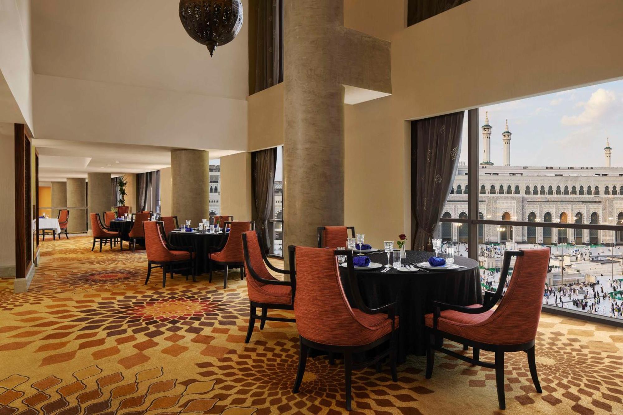 Hilton Suites Jabal Omar Makkah Meca Exterior foto The photo displays the interior of an elegant restaurant. The design features a spacious layout with high ceilings, large windows offering a view of an exterior area, likely a courtyard or plaza. Inside, there are several round tables set with black 