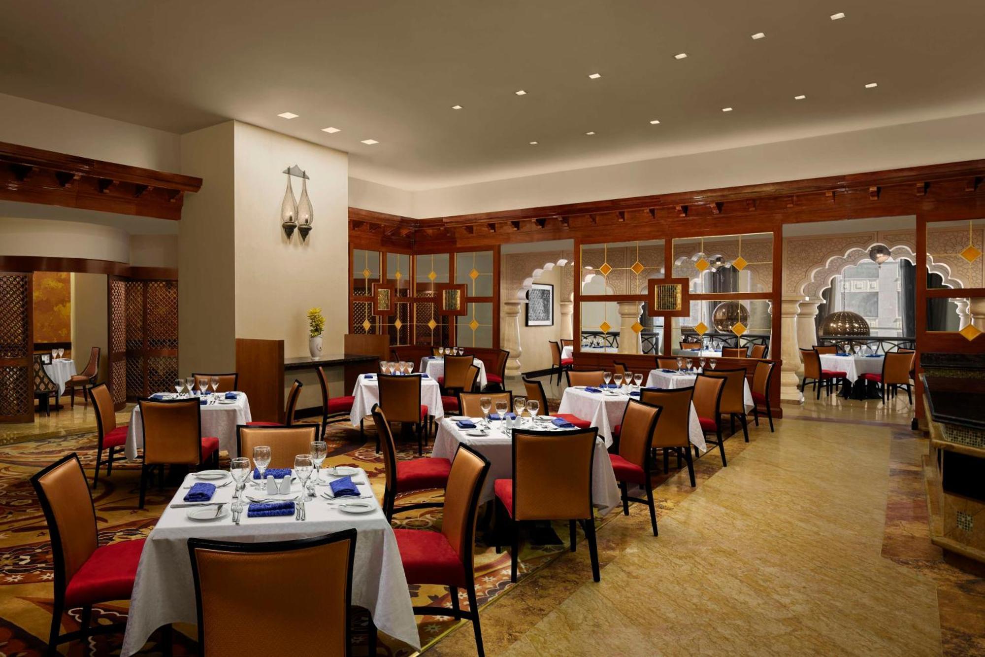 Hilton Suites Jabal Omar Makkah Meca Exterior foto The photo depicts a restaurant interior characterized by elegant decor. The space features several tables set with white tablecloths, plates, cutlery, and blue napkins. The chairs are orange with a contemporary design, and there seems to be a well-li