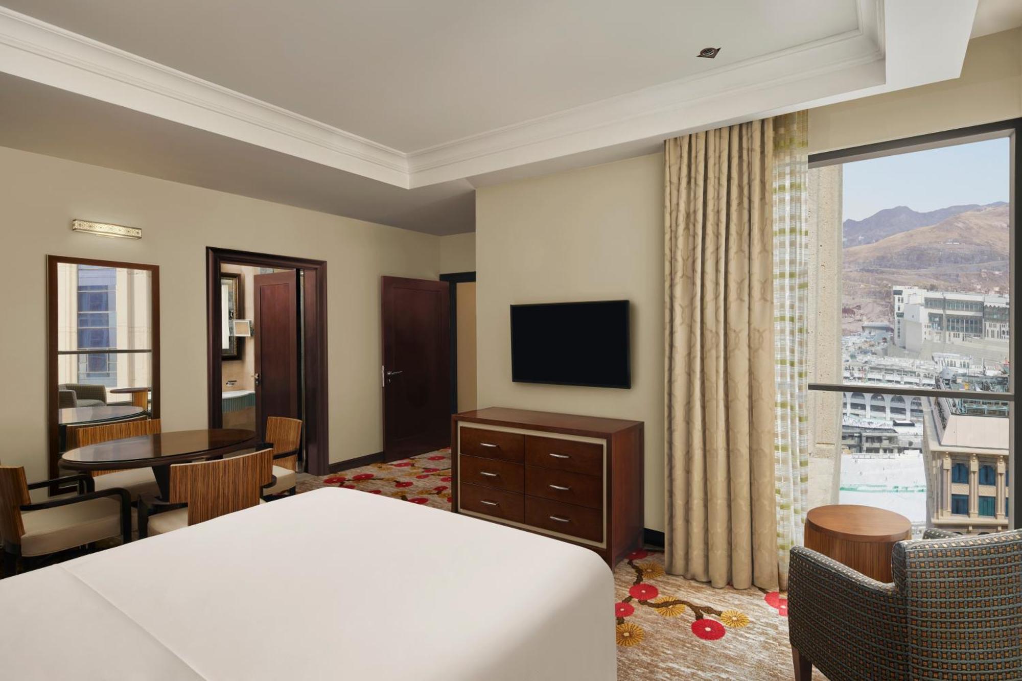 Hilton Suites Jabal Omar Makkah Meca Exterior foto The photo shows the interior of a hotel room. It features a large bed with white bedding in the foreground. In the room, there is a wooden dresser and a flat-screen TV mounted on the wall opposite the bed. To the side, there’s a seating area with a r