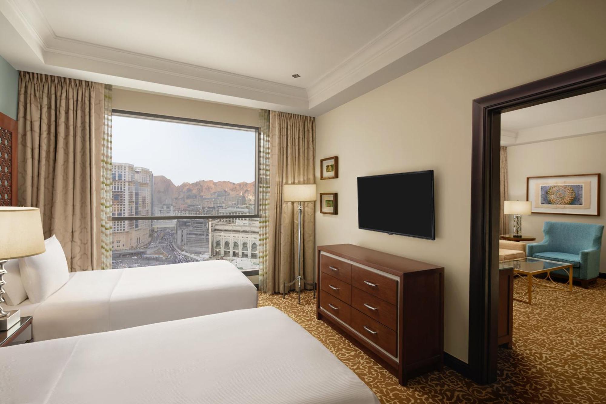 Hilton Suites Jabal Omar Makkah Meca Exterior foto The photo shows a hotel room featuring two beds with white bedding. There is a large window with curtains that allow for natural light, offering a view of the outside scenery, which includes mountains. In the room, there is a wooden dresser and a fla