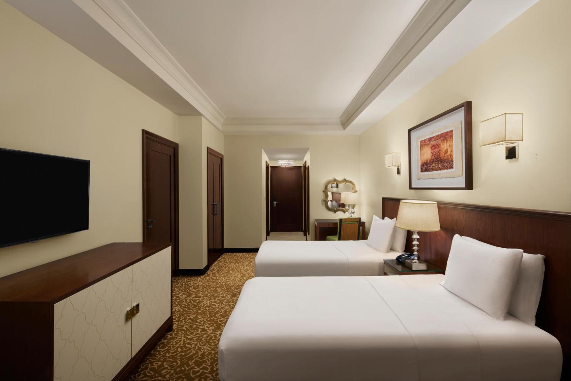 Hilton Suites Jabal Omar Makkah Meca Exterior foto The photo shows a hotel room that features two beds with white linens. The room has light-colored walls and a warm-colored carpet. There is a wooden dresser with a flat-screen TV mounted on the wall opposite the beds. To the side, there are two doors