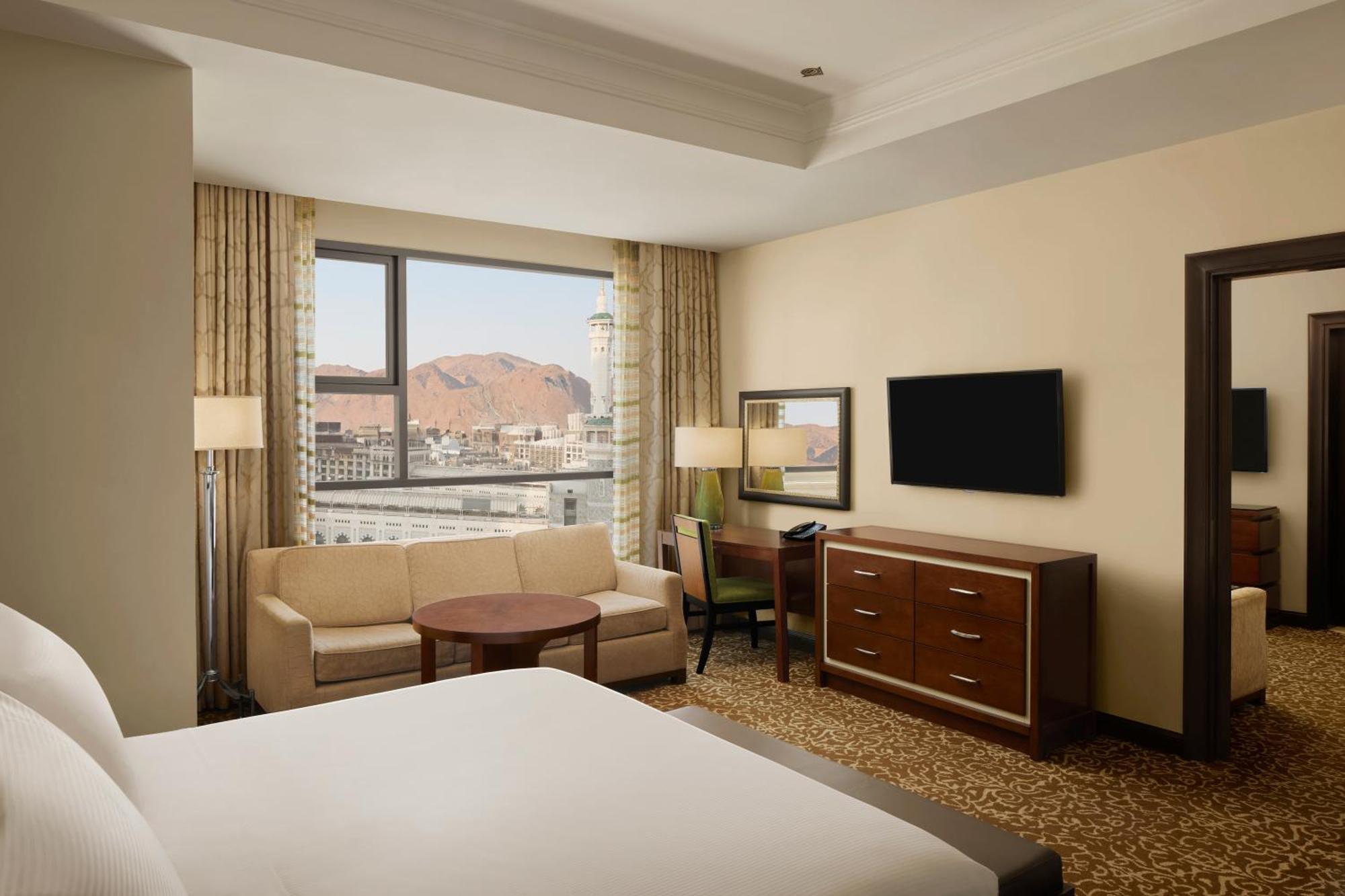 Hilton Suites Jabal Omar Makkah Meca Exterior foto The photo shows a hotel room with modern decor. In the foreground, there is a large bed with white linens. To the left, a comfortable seating area features a sofa, a round coffee table, and a chair. A large window offers a view, likely of a mountain 